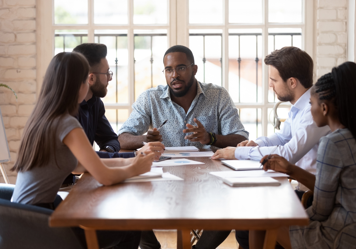 A group of diverse male and female professionals sit at a table in a board room discussing their current talent pipeline.