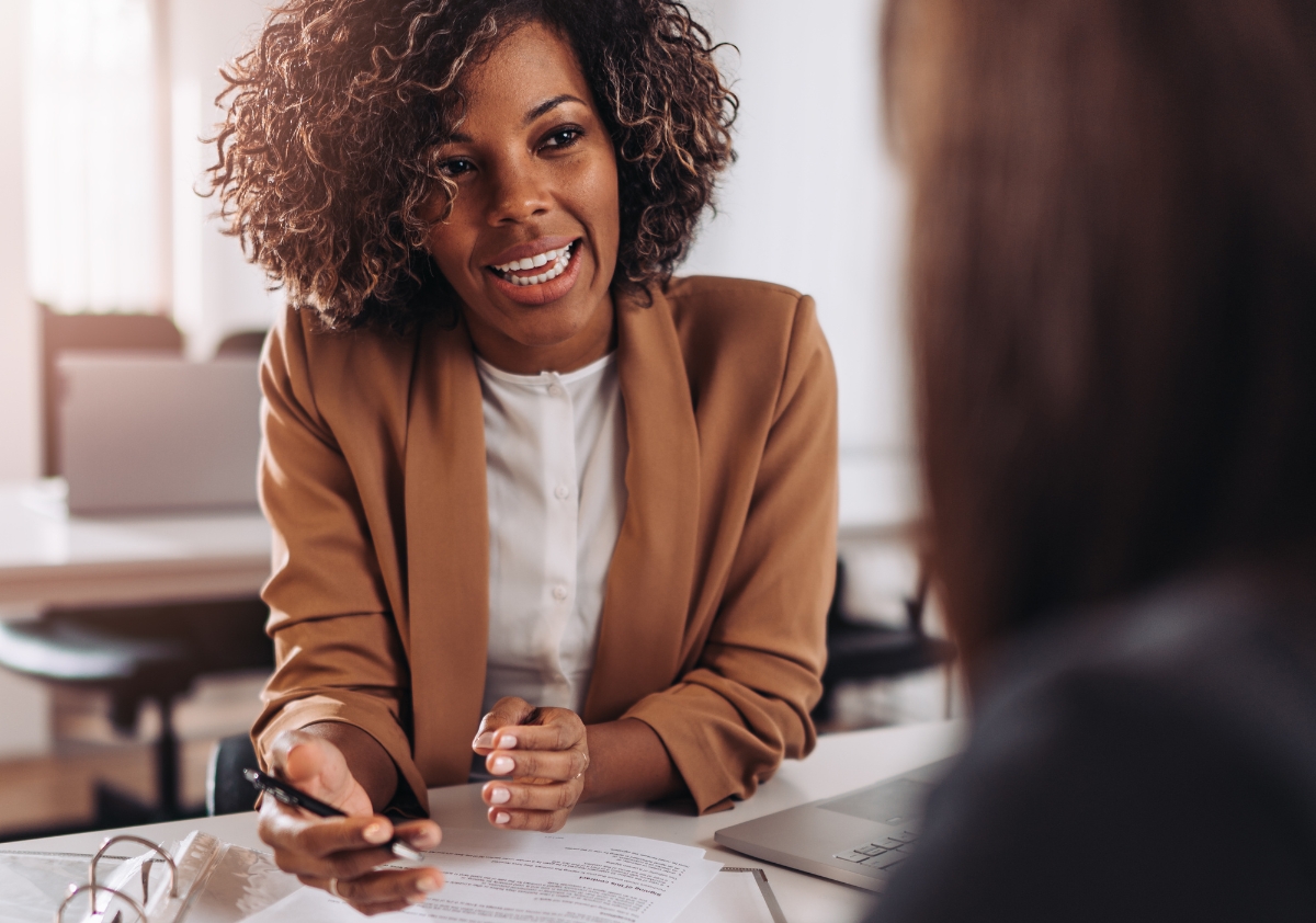 Female Black talent with curly hair wearing a brown blazer and white button up sits at an office desk discussing DEI strategies