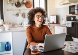 Beautiful Black woman with natural curly hair wearing a burnt orange shirt sits at her kitchen table on her computer reviewing her company's new employee engagement policy