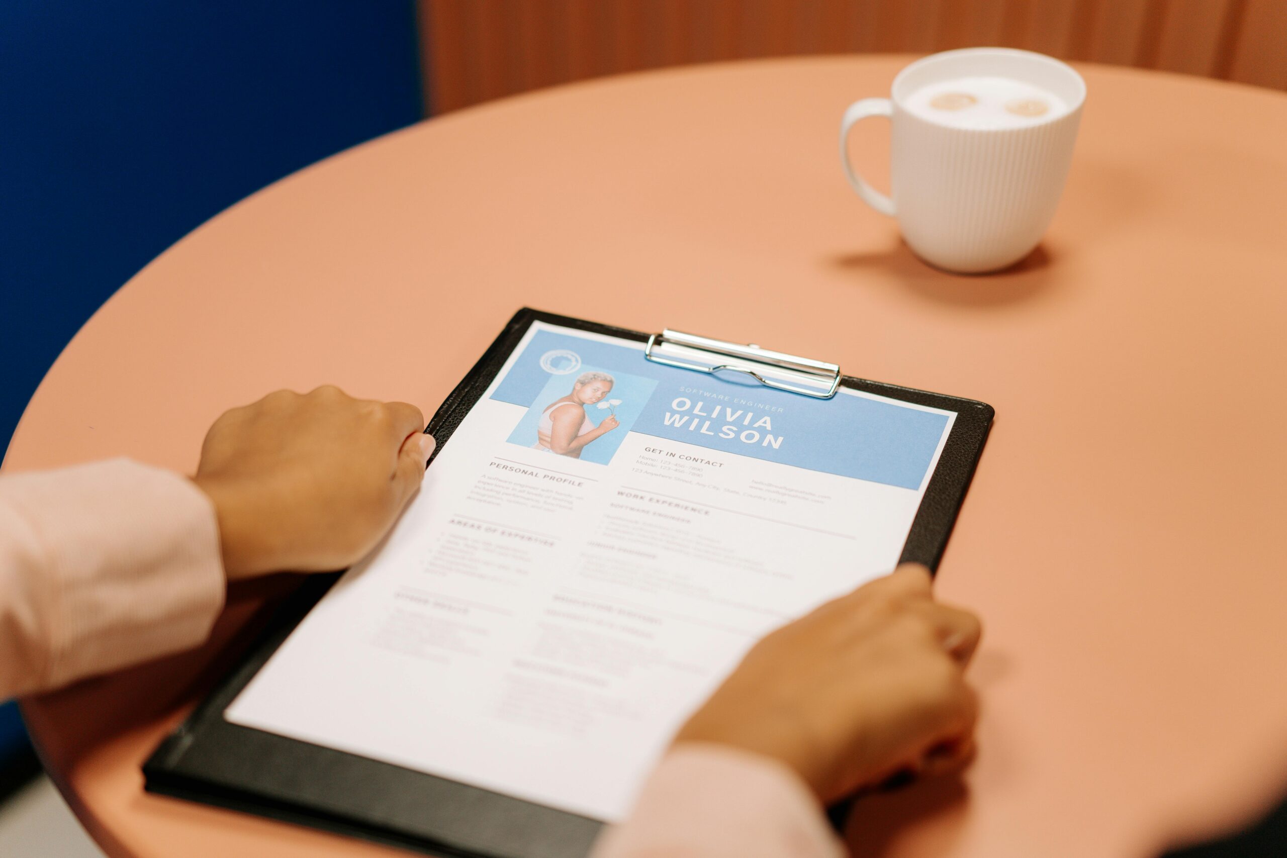 Person holding a clipboard with a resume for 'Olivia Wilson' on a pink table, next to a white cup of coffee, highlighting a job interview or resume review setting.