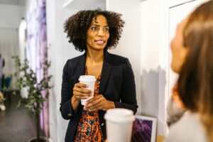 A professional woman with curly hair wearing a navy blazer and a patterned orange dress holds a coffee cup while engaging in conversation with another person out of focus in the foreground. The setting appears to be a casual networking or office environment, with a small plant and wall art in the background.