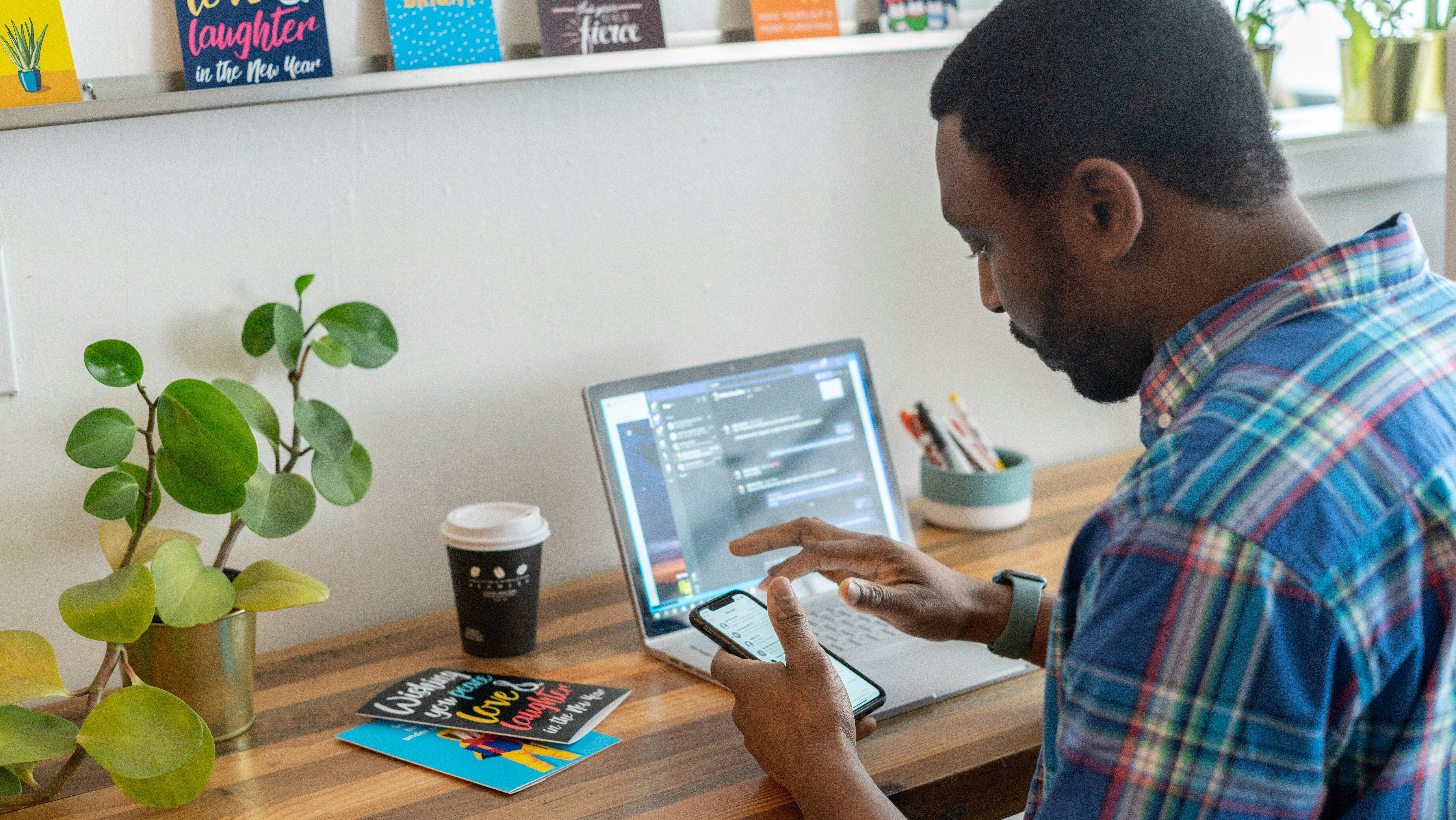 Man sitting at a desk with an open laptop, scrolling through LinkedIn on his phone. How employer branding is a game changer for recruitment.