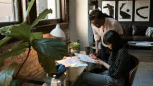 Two women looking at a paper on a clipboard at a desk. Recruitment vs talent acquisition.