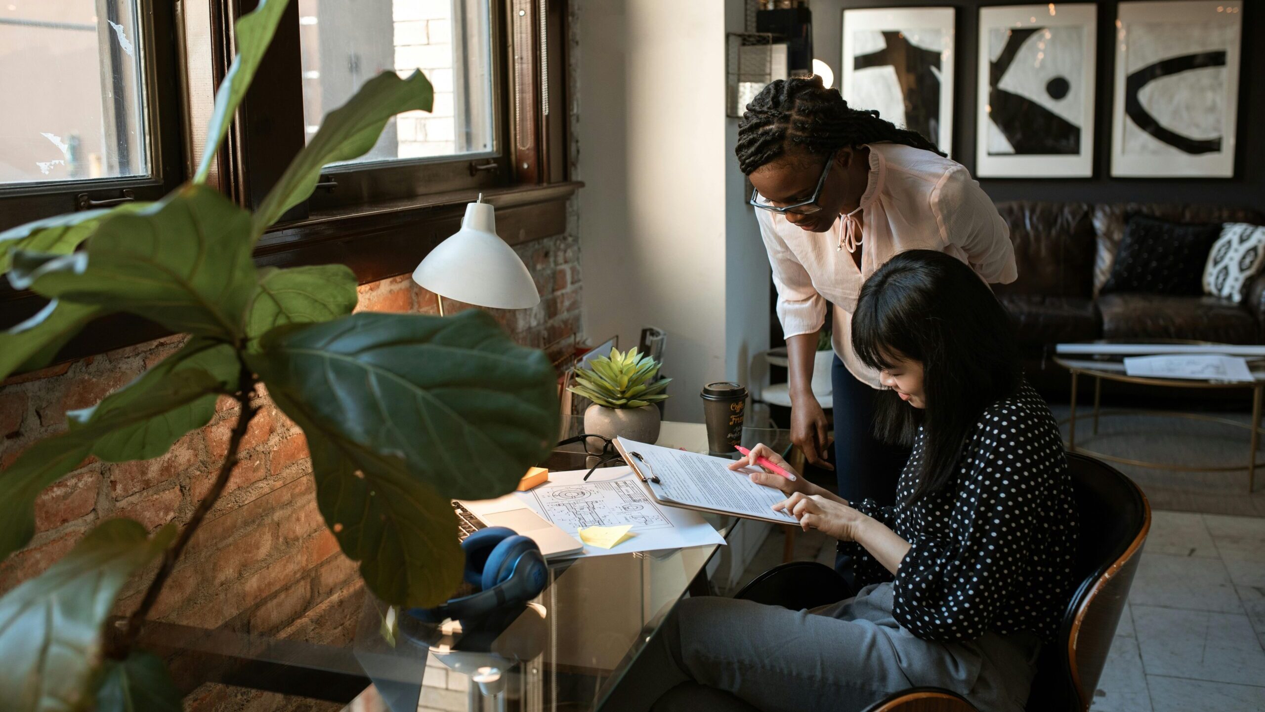 Two women looking at a paper on a clipboard at a desk. Recruitment vs talent acquisition.